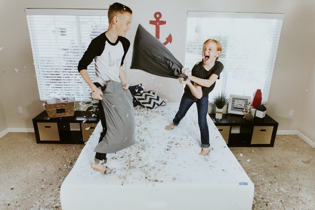 Brother and sister having a pillow fight on a bed with feathers everywhere. If your children’s room is too crowded and you need to create more space, take a look at our simple storage ideas for the kids’ room.